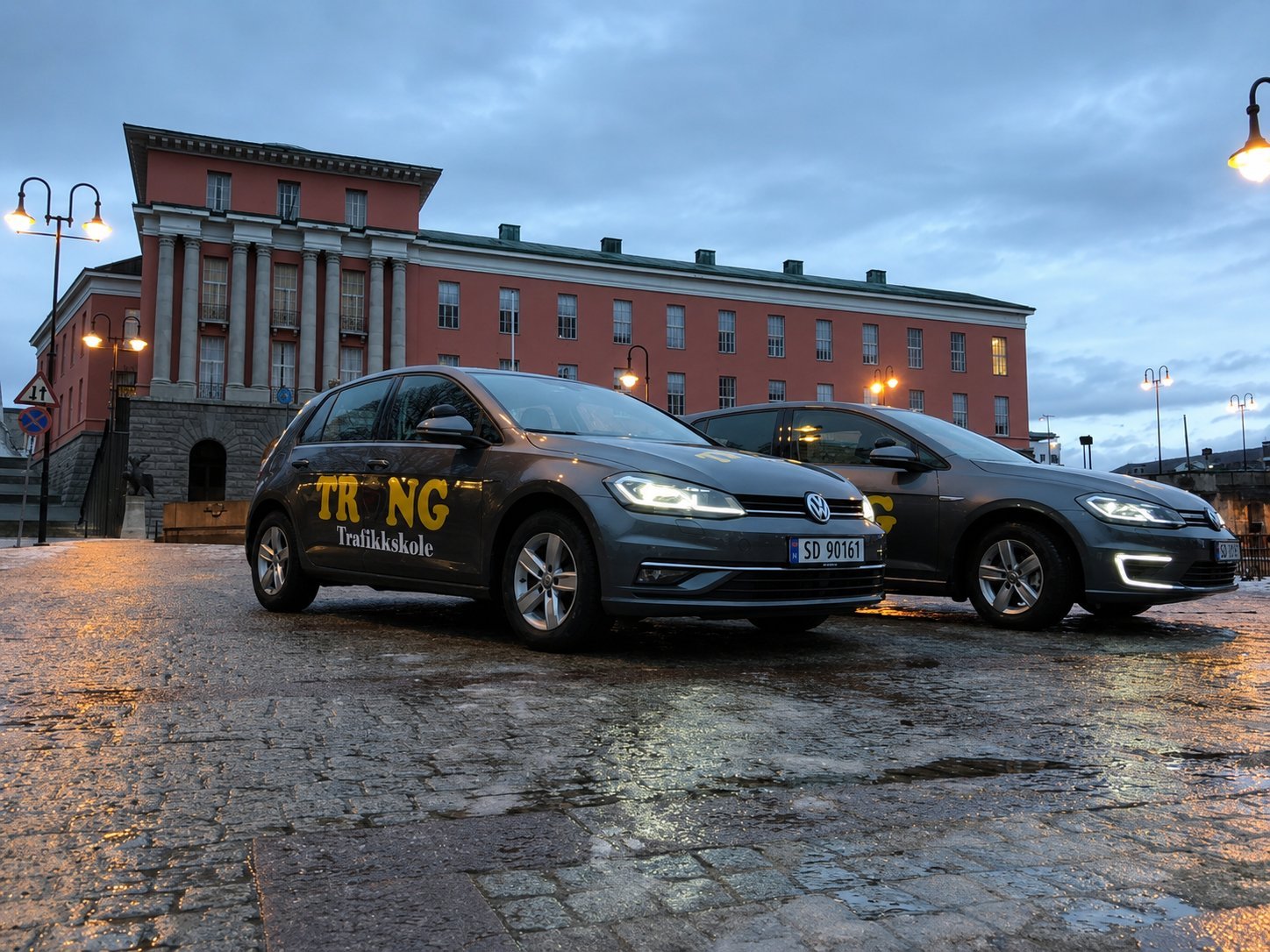 To mørke taxier parkert på våt brosteinsgate ved rød mursteinsbygning i skumring